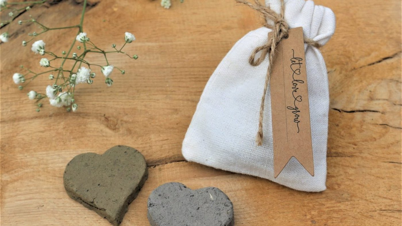 Seed bombs in muted earth tones arranged in a heart shape, with flowers behind